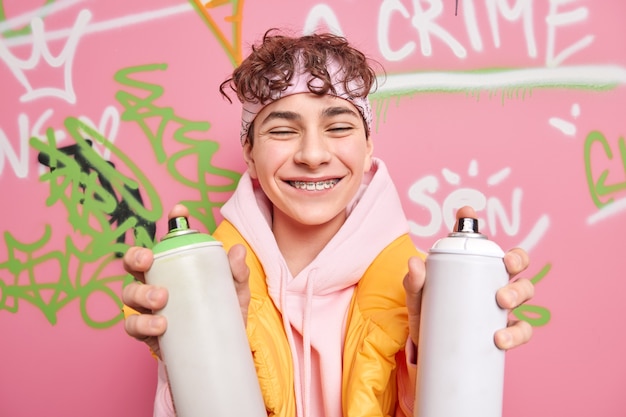 Foto adolescente positivo sonríe con dientes usa aparatos ortopédicos en los dientes cierra los ojos vestido con una sudadera con capucha sostiene dos botellas de spray dibuja graffiti en un lugar público tiene una apariencia fresca disfruta del tiempo libre