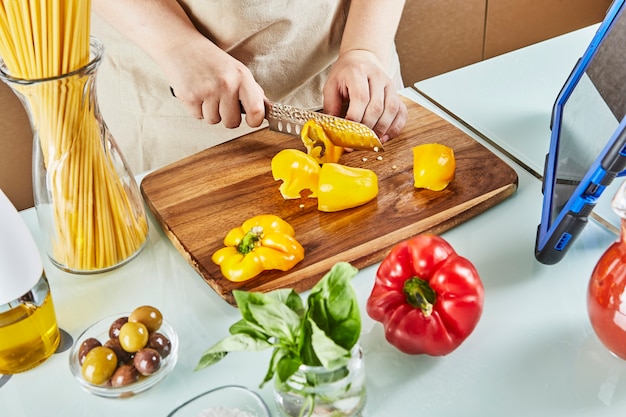 Adolescente prepara un seminario virtual en línea, corta pimiento amarillo, mira una receta digital en una tableta con pantalla táctil mientras prepara una comida saludable en la cocina de su casa.