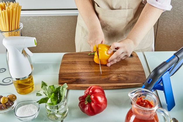 Adolescente prepara un seminario virtual en línea, corta pimiento amarillo, mira una receta digital en una tableta con pantalla táctil mientras prepara una comida saludable en la cocina de su casa.