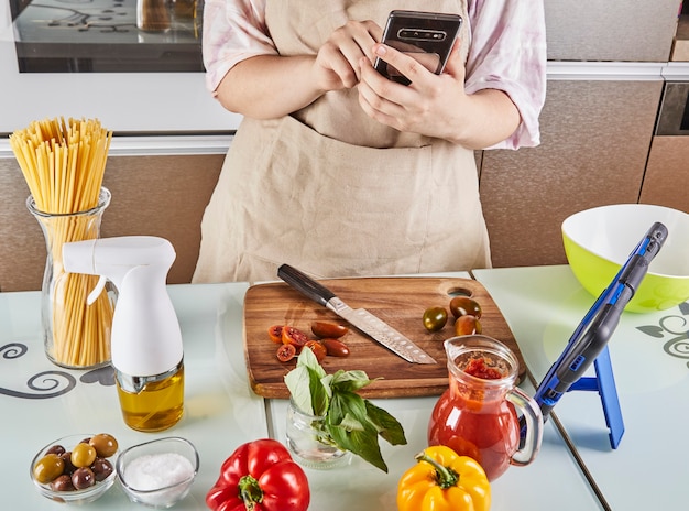 Foto adolescente prepara un tutorial virtual de libros de texto en línea y ve recetas digitales en un teléfono móvil con pantalla táctil mientras prepara alimentos saludables en la cocina de su casa