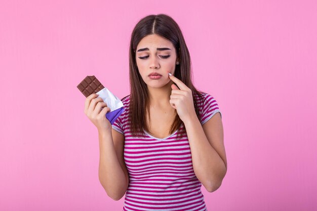 Adolescente con problema de acné sosteniendo una barra de chocolate contra un fondo rosado Joven mujer hermosa Cara de problema de acne con barra de chocolate No feliz comiendo Su cara estresada