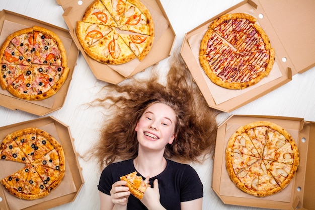 Foto adolescente con una rebanada de pizza en sus manos y cajas de pizza alrededor de sus mentiras