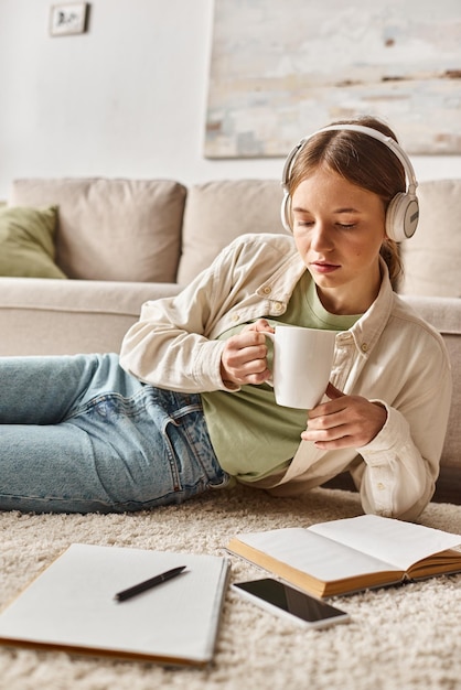 Adolescente relaxada desfrutando de música com fones de ouvido e segurando uma caneca perto de cadernos no tapete