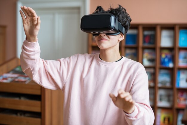 Adolescente en ropa casual mirando la pantalla virtual frente a él mientras realiza una presentación en la biblioteca de la universidad