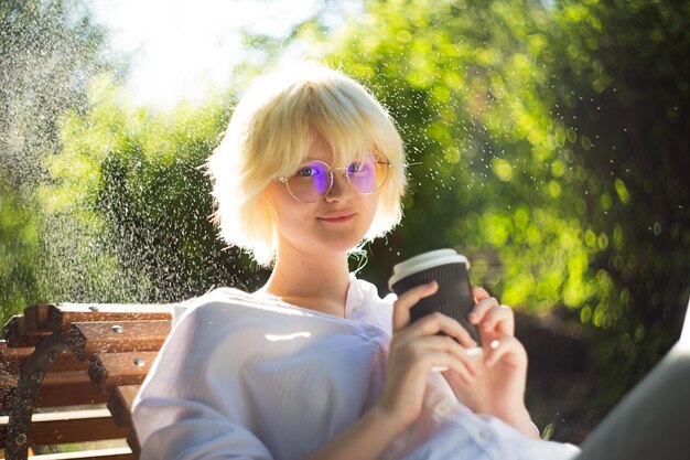 Una adolescente con una rubia escucha beber café de un vaso de papel artesanal afuera