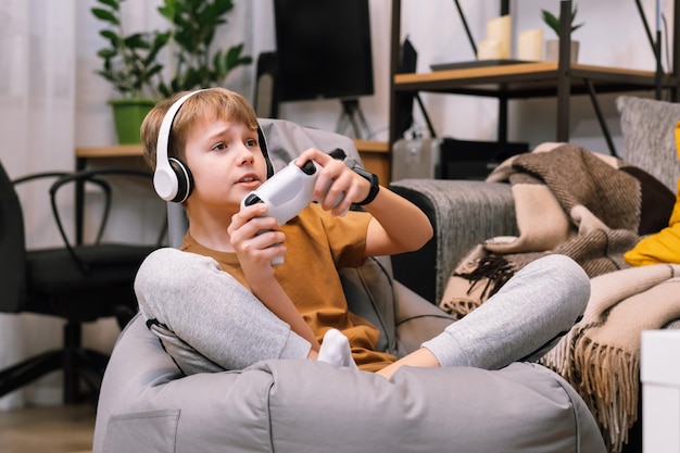 Adolescente rubio con control remoto en la mano jugando a la consola de videojuegos, sentado en una silla sin marco en casa