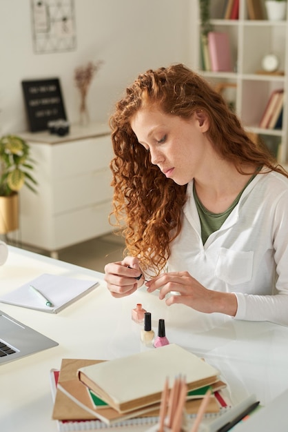 Adolescente ruiva séria com cabelo encaracolado sentada à mesa com cadernos de trabalho e pintando unhas em casa distração