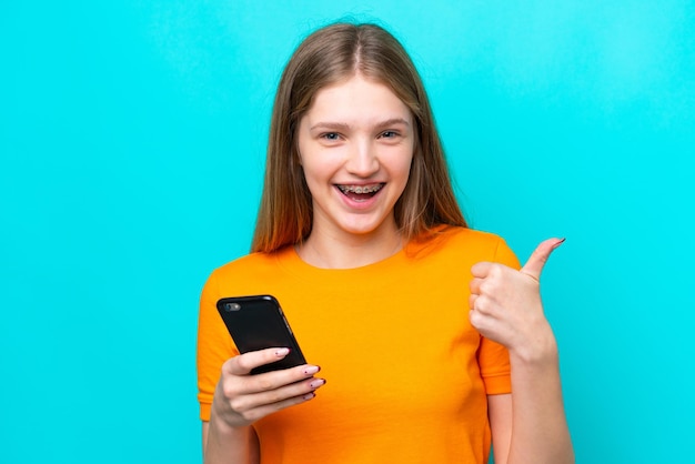 Adolescente rusa aislada de fondo azul a través de un teléfono móvil mientras hace los pulgares hacia arriba