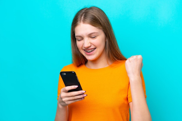 Adolescente rusa aislada de fondo azul usando teléfono móvil y haciendo gesto de victoria