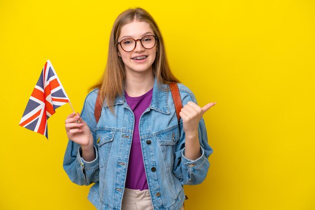 Adolescente rusa sosteniendo una bandera del Reino Unido aislada en un fondo amarillo apuntando hacia un lado para presentar un producto