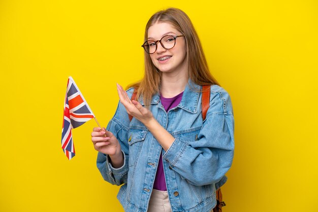 Adolescente rusa sosteniendo una bandera del Reino Unido aislada en un fondo amarillo extendiendo las manos a un lado para invitar a venir