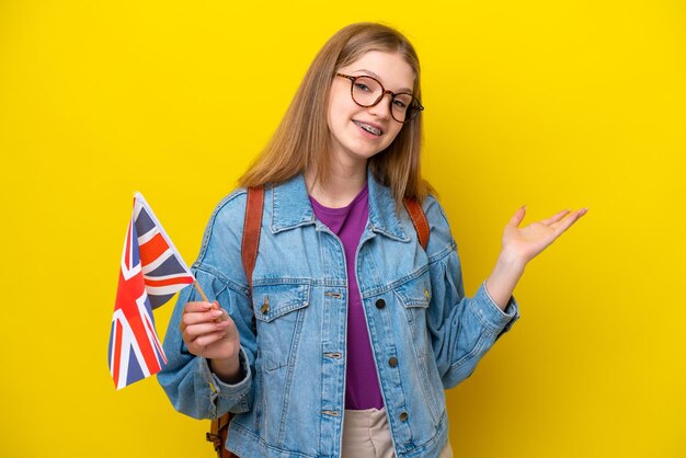 Adolescente rusa sosteniendo una bandera del Reino Unido aislada en un fondo amarillo extendiendo las manos a un lado para invitar a venir