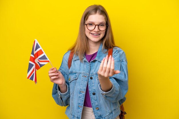 Adolescente rusa sosteniendo una bandera del Reino Unido aislada en un fondo amarillo invitando a venir con la mano Feliz de que hayas venido