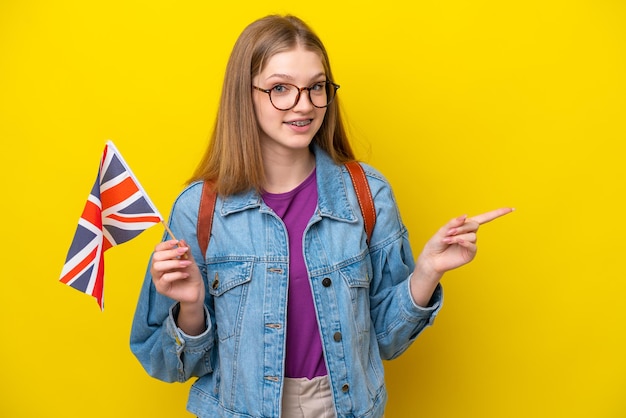 Adolescente rusa sosteniendo una bandera del Reino Unido aislada en un fondo amarillo señalando con el dedo al lado