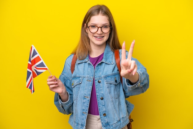 Adolescente rusa sosteniendo una bandera del Reino Unido aislada de fondo amarillo sonriendo y mostrando el signo de la victoria