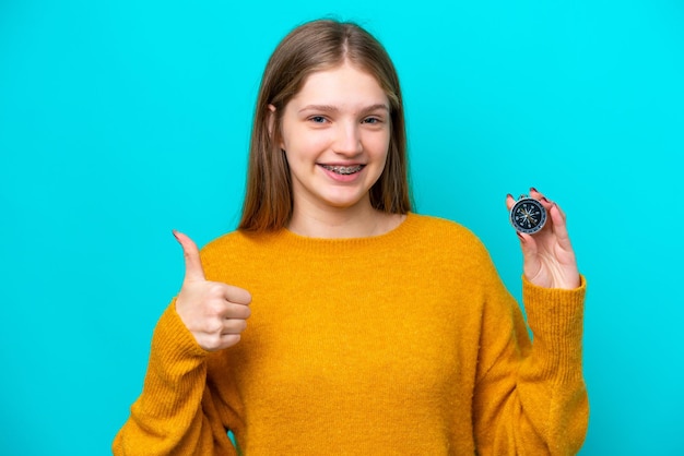 Adolescente rusa sosteniendo una brújula aislada de fondo azul con los pulgares hacia arriba porque algo bueno ha sucedido