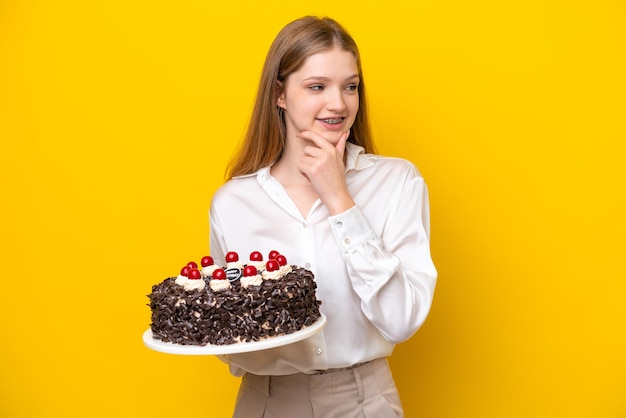 Adolescente rusa sosteniendo pastel de cumpleaños aislado de fondo amarillo mirando hacia un lado y sonriendo