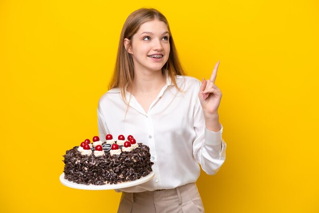 Adolescente rusa sosteniendo pastel de cumpleaños aislado de fondo amarillo señalando una gran idea