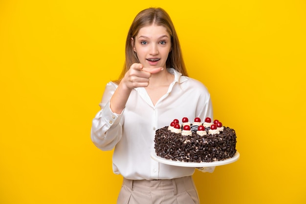 Adolescente rusa sosteniendo pastel de cumpleaños aislado de fondo amarillo sorprendida y apuntando al frente