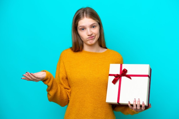 Adolescente rusa sosteniendo un regalo aislado de fondo azul haciendo gestos de duda mientras levanta los hombros