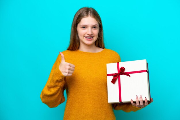 Adolescente rusa sosteniendo un regalo aislado de fondo azul con los pulgares hacia arriba porque algo bueno ha sucedido