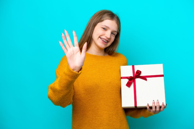 Adolescente rusa sosteniendo un regalo aislado de fondo azul saludando con la mano con expresión feliz