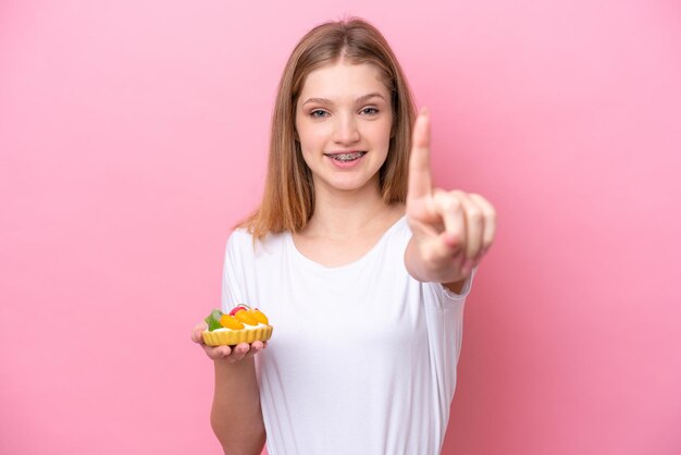 Adolescente rusa sosteniendo una tartaleta aislada de fondo rosa mostrando y levantando un dedo