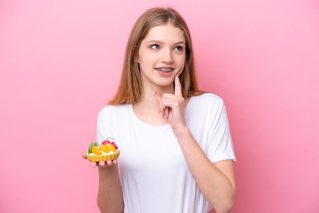 Adolescente rusa sosteniendo una tartaleta aislada de fondo rosa pensando en una idea mientras mira hacia arriba