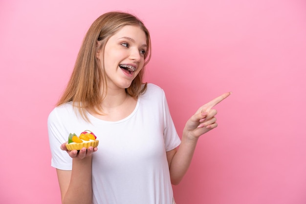 Adolescente rusa sosteniendo una tartaleta aislada de fondo rosa señalando con el dedo al costado y presentando un producto