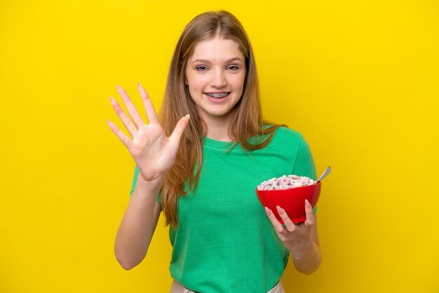 Adolescente rusa sosteniendo un tazón de cereales aislado de fondo amarillo contando cinco con los dedos