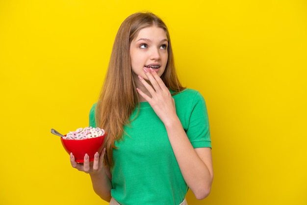 Adolescente rusa sosteniendo un tazón de cereales aislado de fondo amarillo mirando hacia arriba mientras sonríe