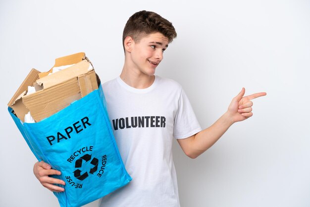 Adolescente ruso hombre sosteniendo una bolsa de reciclaje llena de papel para reciclar aislado sobre fondo blanco apuntando con el dedo al costado y presentando un producto