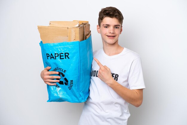 Adolescente ruso hombre sosteniendo una bolsa de reciclaje llena de papel para reciclar aislado sobre fondo blanco apuntando hacia un lado para presentar un producto