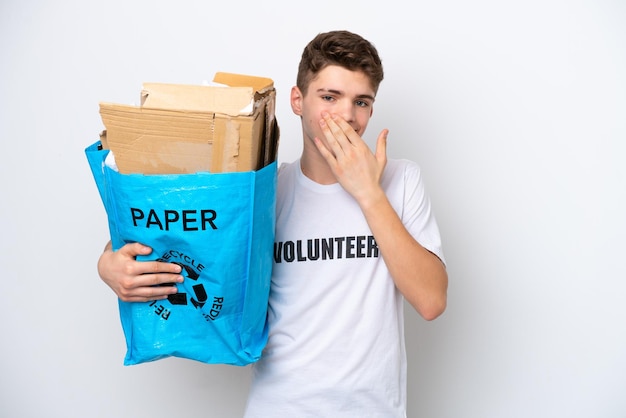 Adolescente ruso hombre sosteniendo una bolsa de reciclaje llena de papel para reciclar aislado sobre fondo blanco feliz y sonriente cubriendo la boca con la mano