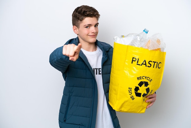 Adolescente russo segurando um saco cheio de garrafas plásticas para reciclar em fundo branco apontando para a frente com expressão feliz