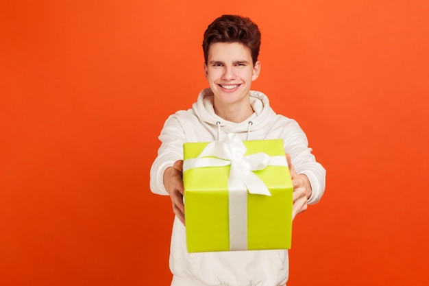 Adolescente satisfeito com capuz branco casual segurando a caixa de presente com sorriso no serviço de entrega de rosto Foto de estúdio interior isolada em fundo laranja