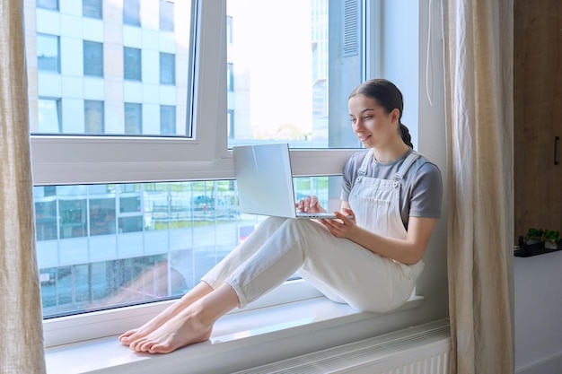 Foto una adolescente sentada en el alféizar de una ventana usando una computadora portátil