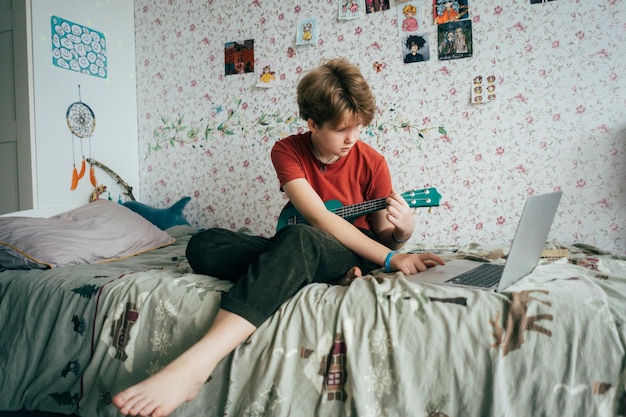 Una adolescente sentada en la cama aprendiendo a tocar el ukulele usando lecciones en línea en una computadora portátil