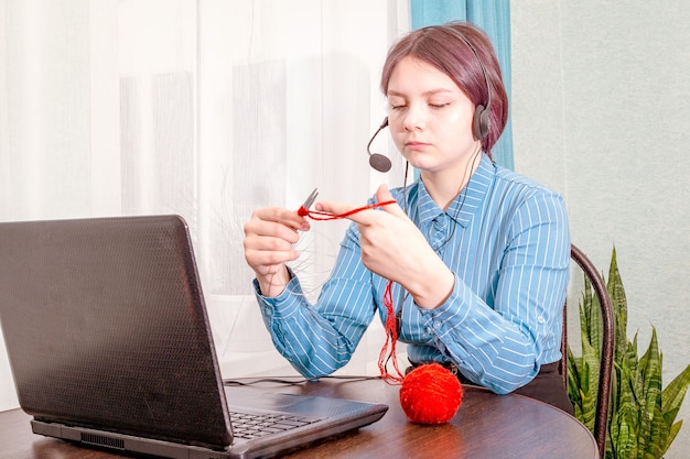 Adolescente sentada frente a su computadora portátil con auriculares, aprendiendo a tejer en línea.