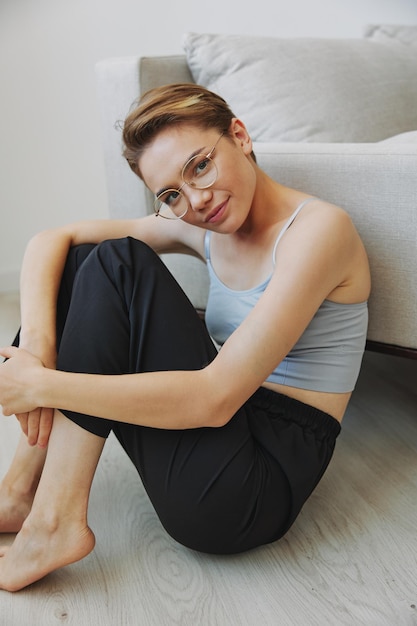 Adolescente sentada no chão em casa sorrindo em roupas de casa e óculos com um estilo de vida de corte de cabelo curto sem filtros espaço de cópia gratuita