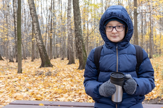 Un adolescente sentado en un banco en el parque de otoño con taza termo llena de bebida caliente, café