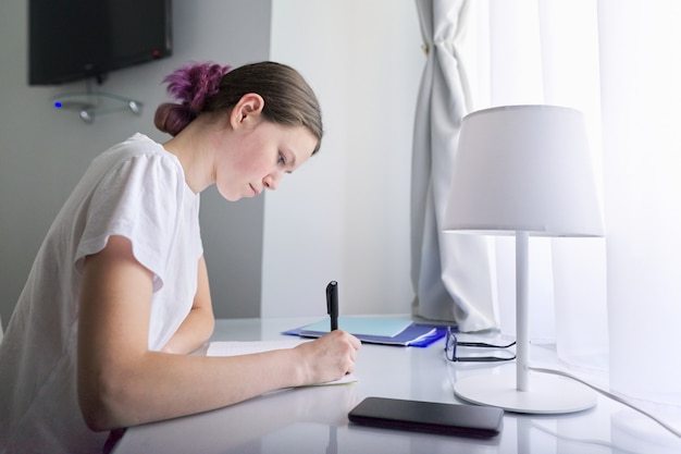 Adolescente sentado en casa en el escritorio cerca de la ventana y escribiendo en el cuaderno