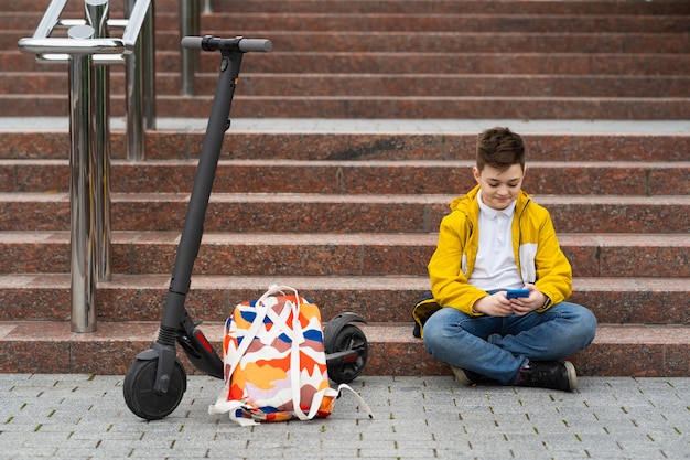 Adolescente sentado cerca de su scooter eléctrico y enviando mensajes de texto en el teléfono inteligente