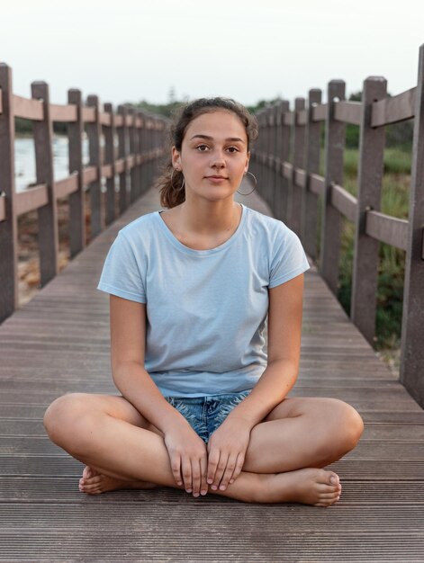 Adolescente sentado de pernas cruzadas na passarela de madeira ao lado do mar