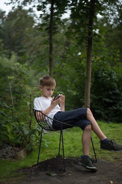 Adolescente sentado em uma cadeira sobre o fundo da natureza e usando um smartphone Quadro vertical