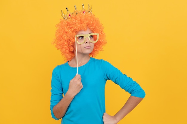 Una adolescente seria con una elegante peluca de payaso usa una corona de reina y divertidas gafas de fiesta, arrogante.