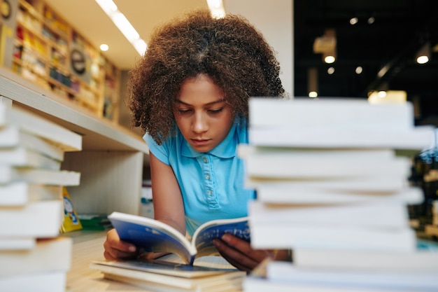 Adolescente séria lendo livro de alunos na biblioteca para um grande projeto escolar