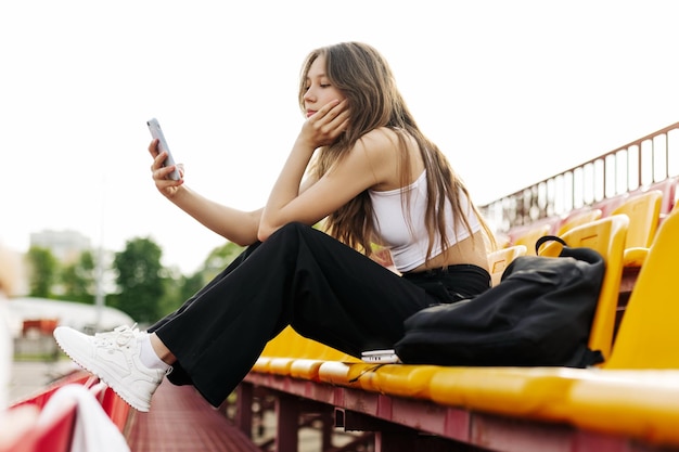 Una adolescente se sienta en las gradas de la escuela y escribe un mensaje en su teléfono en su tiempo libre