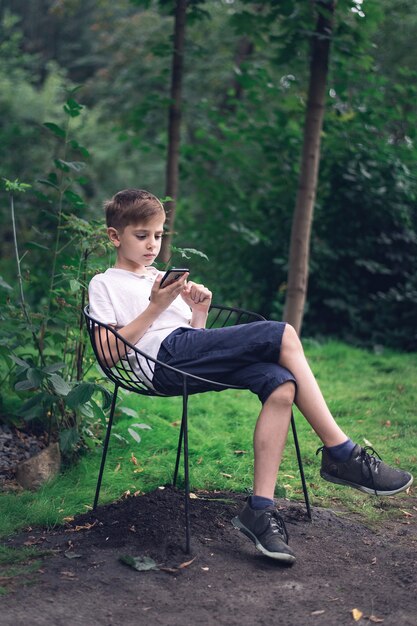 Adolescente se sienta en una silla al aire libre y mirando el teléfono. Marco vertical.