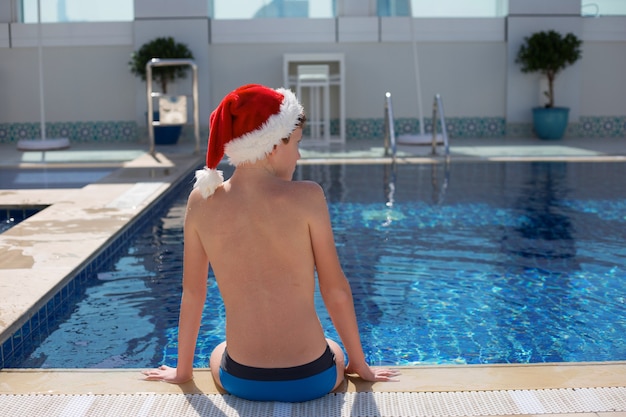 Adolescente con sombrero rojo de Papá Noel sentado en el borde de la piscina en un día soleado